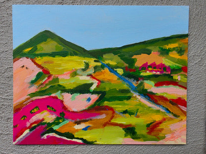 A colorful landscape painting on canvas depicting rolling green hills, a blue river, and vibrant pink fields under a light blue sky, displayed on a textured gray wall.